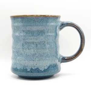 May include: A blue ceramic mug with a textured, ridged surface. The mug has a brown handle and rim. The color gradient transitions from a darker blue at the base to a lighter blue and white near the top. The mug is designed for hot beverages.