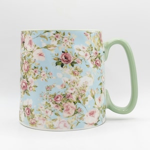 Könnte beinhalten: Keramikbecher mit einem floralen Muster aus rosa und roten Rosen auf hellblauem Hintergrund. Der Becher hat einen gebogenen, hellgrünen Henkel. Geeignet für Tee oder Kaffee.