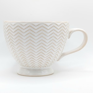 May include: A white ceramic mug with a beige chevron pattern. The mug has a large handle and a pedestal base. The design is repeated around the entire mug. The mug is suitable for coffee, tea, or other beverages.