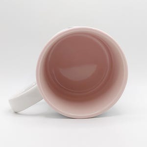 Pode incluir: Uma caneca de cer&acirc;mica branca com interior rosa. A caneca tem uma al&ccedil;a e est&aacute; vazia.
