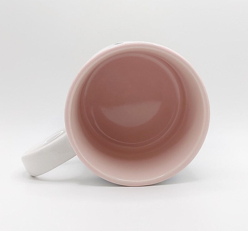 Pode incluir: Uma caneca de cer&acirc;mica branca com interior rosa. A caneca tem uma al&ccedil;a e est&aacute; vazia.