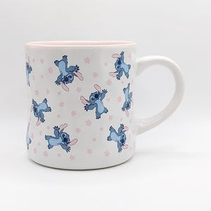 Pode incluir: Caneca de cer&acirc;mica branca com borda e al&ccedil;a rosa. A caneca apresenta um padr&atilde;o repetido de personagens Stitch azuis com flores rosas em um fundo branco.