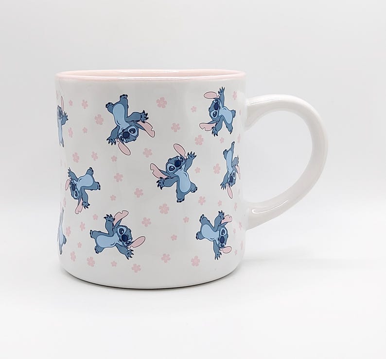 Pode incluir: Caneca de cer&acirc;mica branca com borda e al&ccedil;a rosa. A caneca apresenta um padr&atilde;o repetido de personagens Stitch azuis com flores rosas em um fundo branco.