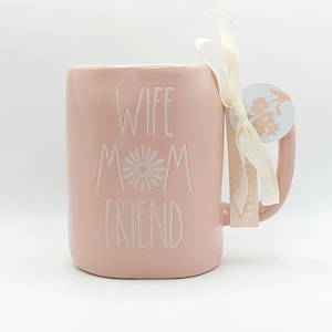 Op de afbeelding: Een roze keramische mok met de tekst "Wife Mom Friend" in witte letters. De mok heeft een wit bloemenontwerp en een wit lint om de handgreep.