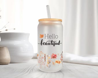 May include: Frosted glass can-shaped tumbler with a bamboo lid and a metal straw. The tumbler is decorated with the words "Hello beautiful" and floral illustrations. The design features orange, yellow, and red flowers and foliage.