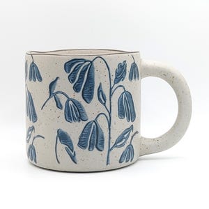 May include: A ceramic mug with a speckled off-white finish and a blue botanical pattern featuring drooping flowers and leaves. The mug has a thick, rounded handle and a slightly uneven, organic rim, showcasing a hand-crafted appearance.