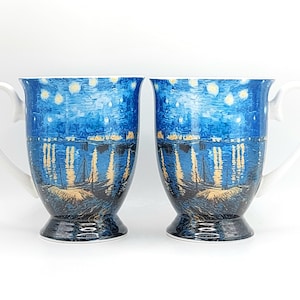 May include: Two white-handled coffee mugs with a blue and yellow night scene design. The design depicts a starry night sky over a body of water with boats and buildings. The mugs have a pedestal base.
