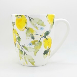 May include: White ceramic mug with a watercolor design of yellow lemons, green leaves, and small blue flowers. The mug has a curved handle and a smooth, glossy finish. The design wraps around the mug.