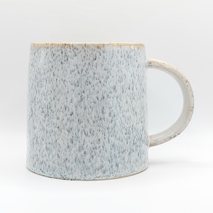 Könnte beinhalten: Eine Keramiktasse mit einer gesprenkelten, hellblauen und weißen Glasur. Die Tasse hat einen abgerundeten Henkel und einen hellbraunen Rand. Die Tasse ist zylindrisch und steht auf einer weißen Oberfläche.