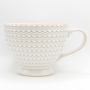 May include: Cream-colored ceramic mug with a scalloped design. The mug has a large handle and sits on a small pedestal base. The exterior features horizontal rows of wave-like patterns.