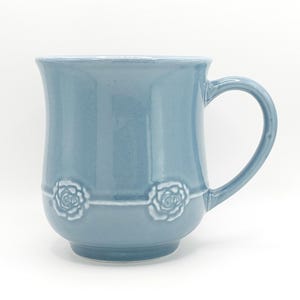May include: A light blue ceramic mug with a curved handle. The mug features a decorative band with two embossed rose designs. The mug has a slightly flared top and a rounded base, suitable for hot or cold beverages.