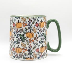 May include: A white ceramic mug with a green rim and handle, featuring a repeating pattern of orange pumpkins, red apples, tan oak leaves, and small blue and purple floral sprigs scattered across the exterior surface of the cylindrical vessel.