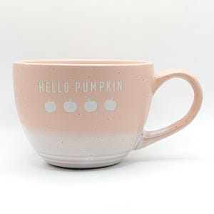 Könnte beinhalten: Eine rosafarbene Keramiktasse mit weißem Rand und Henkel. Die Tasse hat eine gesprenkelte Oberfläche und trägt die Aufschrift "HELLO PUMPKIN" in weißen Buchstaben mit vier weißen Kürbis-Silhouetten.