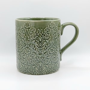 May include: A sage green ceramic mug featuring a raised, embossed floral and botanical pattern covering the entire exterior surface. The mug has a smooth, glossy finish, a cylindrical body, and a curved handle on the right side.