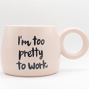 May include: A blush pink ceramic mug with a rounded body and a circular handle. The mug features the phrase "I'm too pretty to work" in black script. The mug is designed for beverages.