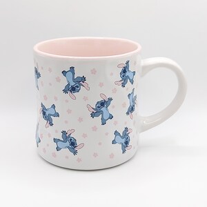 Pode incluir: Caneca de cer&acirc;mica branca com uma borda rosa e um padr&atilde;o repetido de personagens Stitch azuis do filme da Disney Lilo & Stitch. Os personagens Stitch est&atilde;o em uma pose brincalhona com os bra&ccedil;os estendidos.