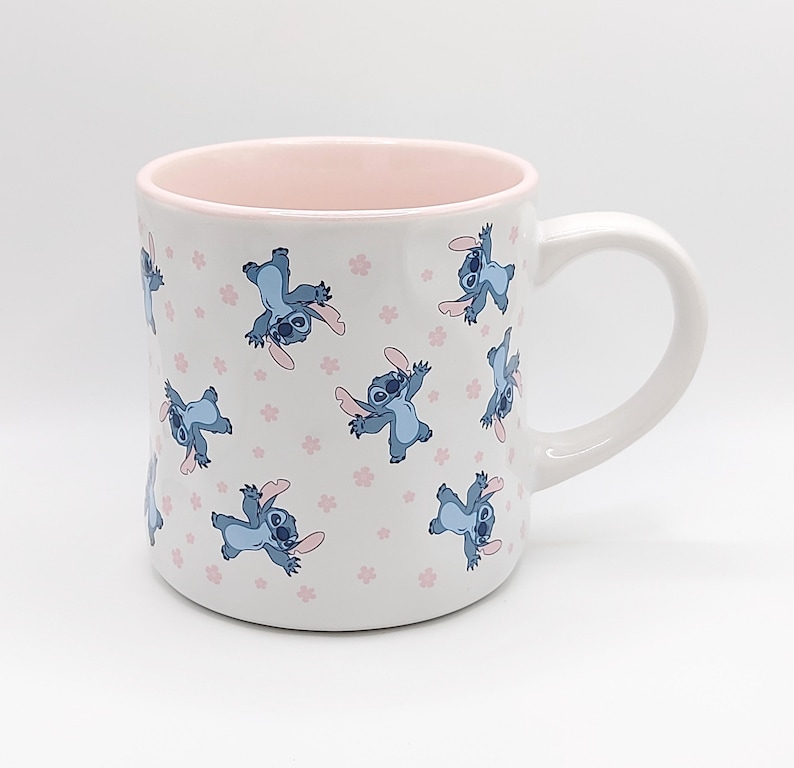 Pode incluir: Caneca de cer&acirc;mica branca com uma borda rosa e um padr&atilde;o repetido de personagens Stitch azuis do filme da Disney Lilo & Stitch. Os personagens Stitch est&atilde;o em uma pose brincalhona com os bra&ccedil;os estendidos.