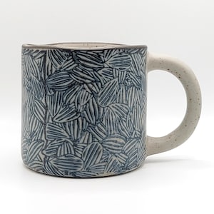 May include: A ceramic mug with a speckled off-white base and a dark blue hand-carved leaf pattern covering the exterior. The mug features a sturdy, matching speckled handle and a smooth, rounded rim, showcasing a textured, artisanal finish.