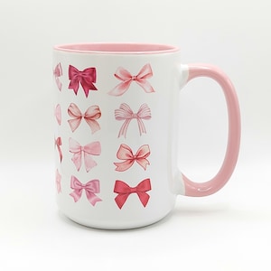 May include: White ceramic mug with a pink rim and handle. The mug features a pattern of pink bows in various shades and sizes.