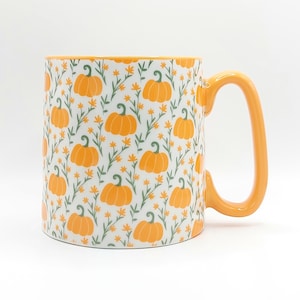 Op de afbeelding: Een witte keramische mok met een oranje rand en handvat. De mok is versierd met een patroon van oranje pompoenen en groene bladeren op een witte achtergrond.