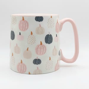 May include: White ceramic mug with a pink handle and rim. The mug is decorated with a pattern of pumpkins in shades of pink, gray, and beige. The pumpkins have various designs, including stripes and dots.