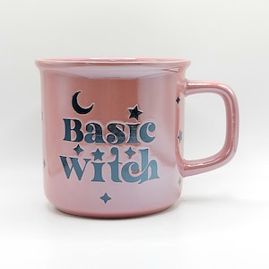 Puede incluir: Una taza de cerámica de color rosa claro brillante con el texto azul oscuro "Basic witch" impreso en la parte frontal. El diseño incluye una luna creciente y varias estrellas pequeñas de cuatro puntas dispersas alrededor de las letras.