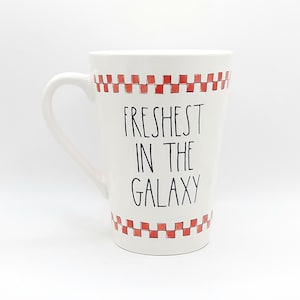 May include: White ceramic mug with a curved handle. The mug features the text "Freshest in the Galaxy" in black, with a red and white checkered pattern at the top and bottom. The mug is designed for hot beverages.