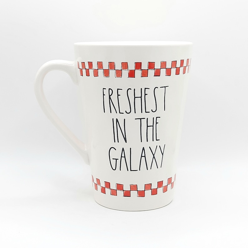 May include: White ceramic mug with a curved handle. The mug features the text "Freshest in the Galaxy" in black, with a red and white checkered pattern at the top and bottom. The mug is designed for hot beverages.