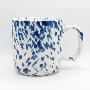 May include: A ceramic mug with a white base and a blue speckled pattern. The mug features a cylindrical body and a curved handle on the right side. The blue paint appears as various sized dots and splatters distributed across the entire surface.