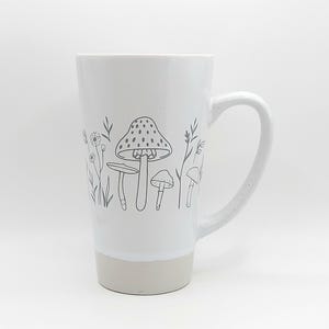 May include: White ceramic mug with a gray outline of mushrooms and flowers. The mug has a handle and a light gray band around the bottom.