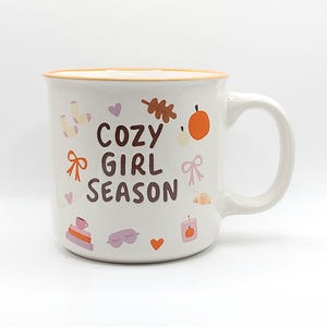 May include: A white ceramic mug with an orange rim featuring the brown text COZY GIRL SEASON. The design includes small illustrations of socks, hearts, an oak leaf, a pumpkin, bows, a croissant, a stack of books with a cup, a sleep mask, and a candle.