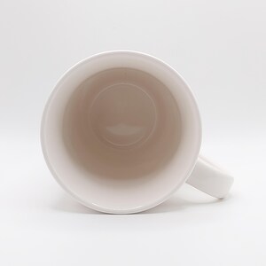 May include: A white ceramic mug with a rounded interior and a handle. The inside of the mug is a light beige color, and the outside is white. The mug is set against a white background.