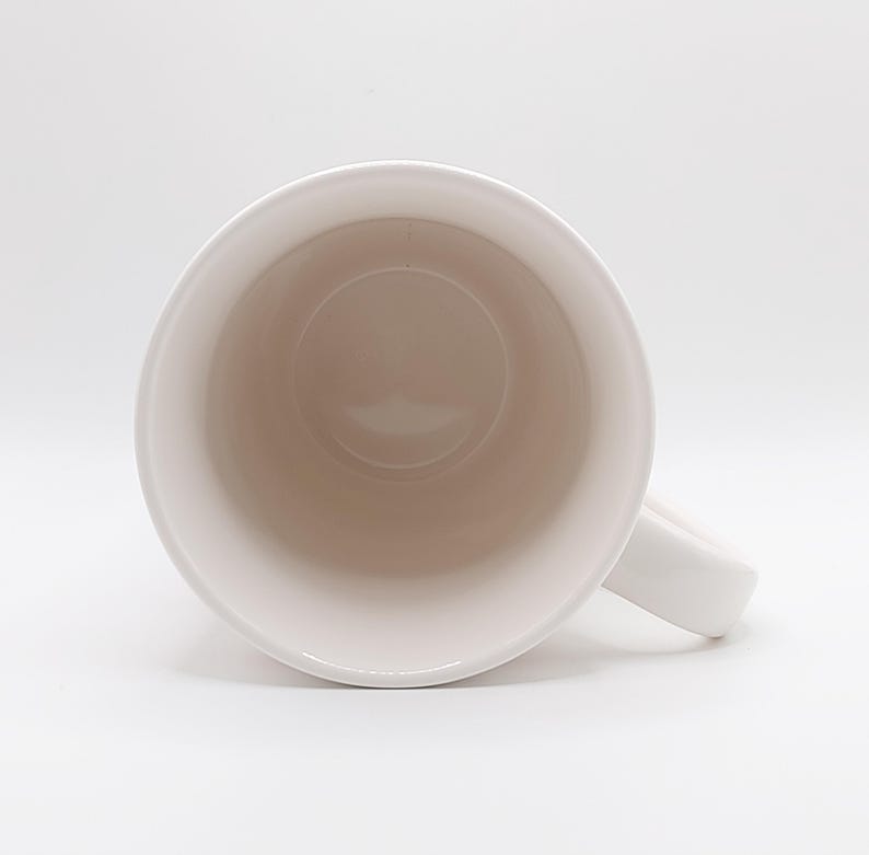 May include: A white ceramic mug with a rounded interior and a handle. The inside of the mug is a light beige color, and the outside is white. The mug is set against a white background.