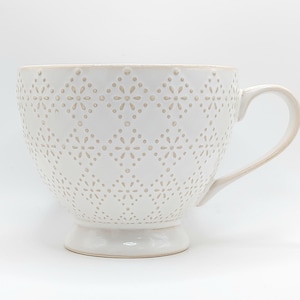 May include: A white ceramic teacup with a decorative floral pattern and a handle. The teacup has a wide bowl shape and sits on a small pedestal base. The teacup is approximately 5 inches in diameter and 4 inches tall.