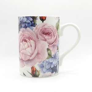 May include: White ceramic mug featuring a floral design with large pink roses, blue hydrangeas, and orange buds. The mug has a curved handle and a white background. The floral pattern covers the majority of the mug's surface.