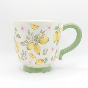 May include: A white ceramic mug with a green handle and base. The mug is decorated with yellow lemons, pink flowers, and green leaves. The mug is designed for hot beverages.