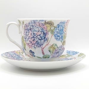 May include: A white porcelain teacup and saucer set. The teacup and saucer are decorated with a floral hydrangea pattern in blue, pink, and purple. The teacup has a curved handle and sits on the matching saucer.