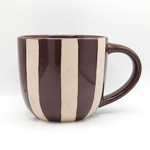 May include: A ceramic mug featuring a glossy dark brown finish with vertical cream-colored stripes. The cream stripes have a speckled texture throughout, while the dark brown sections are smooth and solid. The mug includes a curved handle on the right.