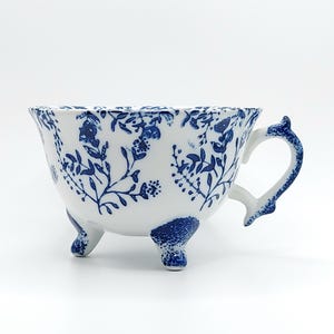 May include: A white ceramic teacup featuring a blue floral pattern across the exterior and handle. The cup is supported by three small, blue-patterned feet, with the blue design extending onto the base of each foot and the curved handle.