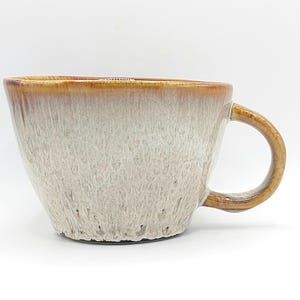 Könnte beinhalten: Eine Keramik-Kaffeetasse mit einem einzigartigen Design. Die Tasse hat eine hellbeige Außenseite mit strukturierter Optik und einen braunen Rand und Henkel. Der Henkel ist rund und die Tasse hat eine weite Öffnung.