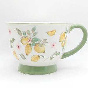 May include: White ceramic mug with a green handle and base, adorned with watercolour lemons, pink flowers, and green leaves. The mug has a wide, rounded shape, ideal for a warm drink. A charming addition to any kitchen, with a fresh, summery design.