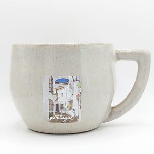 May include: A speckled, off-white ceramic mug with a rounded body and a large handle. A sticker on the side features a photo of a street scene. The mug is labeled "Made in Portugal".