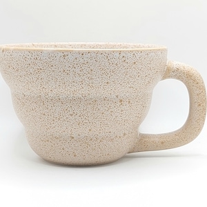 May include: A speckled beige ceramic mug with a rounded handle. The mug has a wide, slightly flared rim and a textured surface. The mug's design features a series of horizontal ridges around the body.