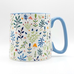 May include: A ceramic mug with a white background, adorned with a colorful floral pattern of leaves, mushrooms, and berries. The mug has a light blue handle and rim, perfect for enjoying your favorite beverage.