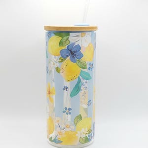 May include: A clear glass tumbler with a bamboo lid and a clear straw. The tumbler is decorated with a lemon and floral design on a light blue striped background. The design includes yellow lemons, blue and white flowers, and green leaves.