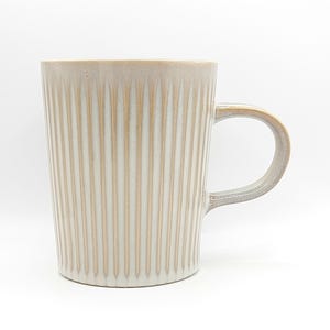 May include: A beige ceramic mug with vertical ribbed detailing. The mug has a rounded handle and a neutral color palette, suitable for coffee or tea. The mug is approximately 5 inches tall.
