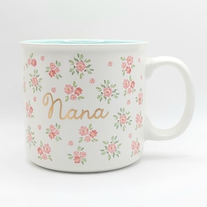 May include: White ceramic mug with a floral pattern of pink flowers and green leaves. The word "Nana" is written in gold script. The inside of the mug is a light blue color.
