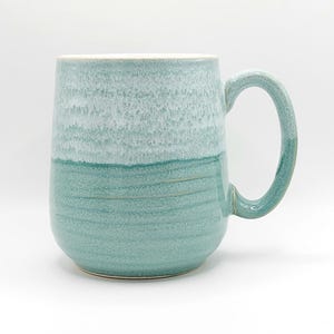 Może przedstawiać: Duży, dwutonowy ceramiczny kubek z zaokrągloną podstawą i dużym uchwytem. Górna połowa kubka jest jasna, teksturowana niebiesko-zielona, przechodząca w ciemniejszą, gładką niebiesko-zieloną w dolnej połowie. Do gorących napojów.