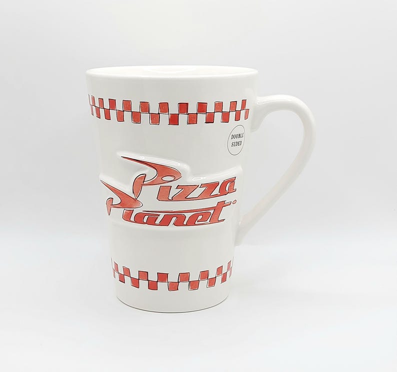 May include: White ceramic mug with a large handle, featuring the Pizza Planet logo in red. The mug has a red and white checkered pattern around the top and bottom. The words "DOUBLE SIDED" are printed on the side.