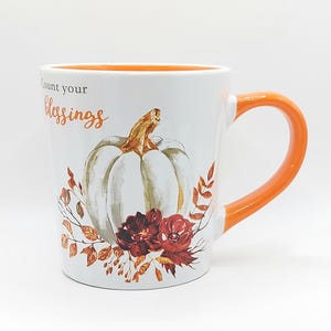 May include: White ceramic mug with an orange handle and interior. The mug features a watercolor design of a white pumpkin, burgundy flowers, and fall foliage. The text "Count your blessings" is written on the mug.